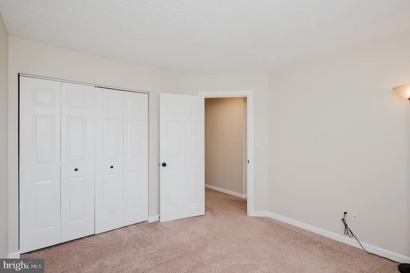 Empty beige bedroom with light brown carpet, a double closet with white doors on the left, and an open doorway to a hallway on the right; a wall sconce provides light.