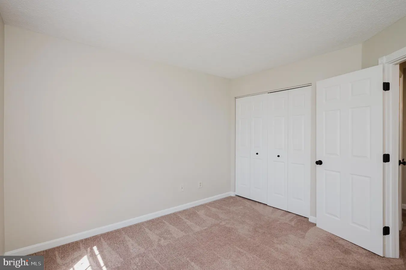 Empty beige bedroom with light carpet, white closet doors, and an open entry door to another room.