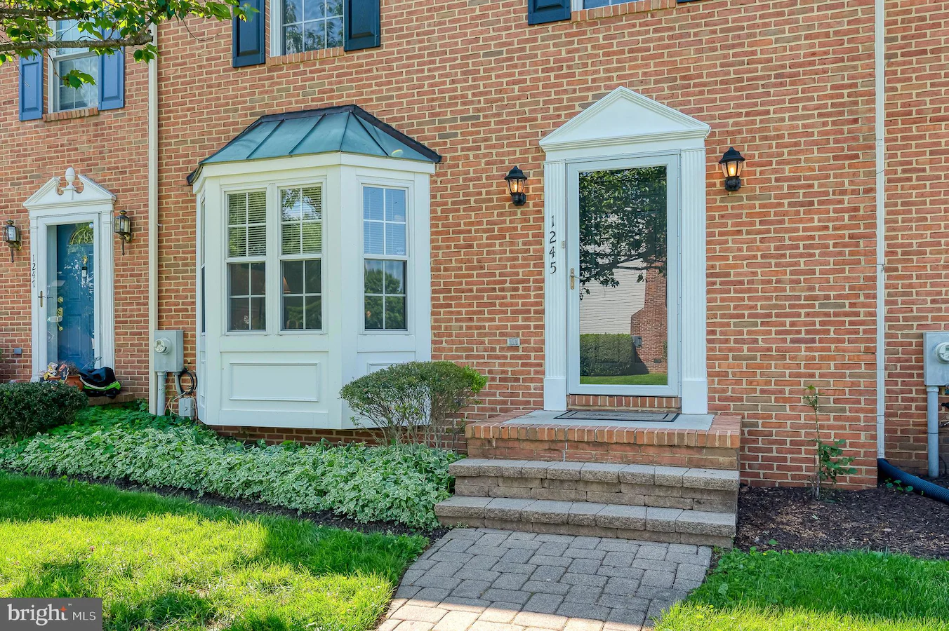 Brick townhouse facade with three entrances: a blue door on the left, a white bay-windowed nook in the center, and a glass door on the right, with a manicured lawn and shrubs nearby.