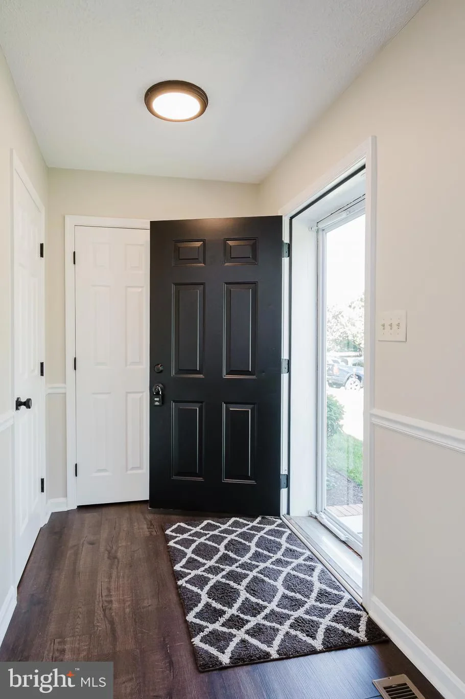 Open black front door in a bright entry hall with white doors, dark wood floor, and a gray geometric rug by the doorway.