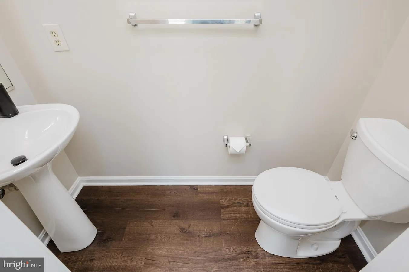 Small bathroom with a white pedestal sink on the left, white toilet on the right, chrome towel bar, toilet paper holder, and dark wood-look flooring.