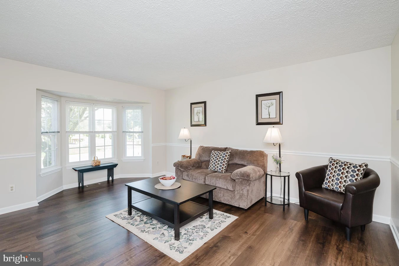 Living room with beige sofa, brown leather chair, coffee table, and bay window area in a bright, neutral decor.