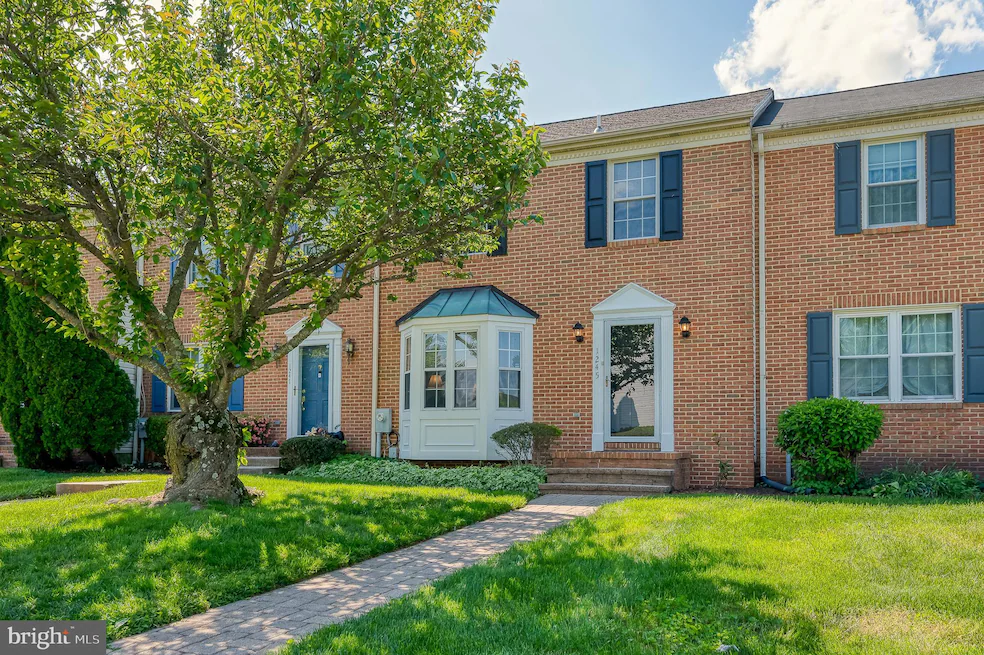 Brick townhouse with blue shutters and a white bay window, set in a green front yard with a large tree and brick walkway leading to the front door.