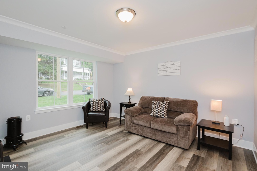Bright living room with light gray walls, a brown sofa with patterned cushions, a black armchair by a window, two dark wood side tables with lamps, and a small wall art piece; light wood flooring