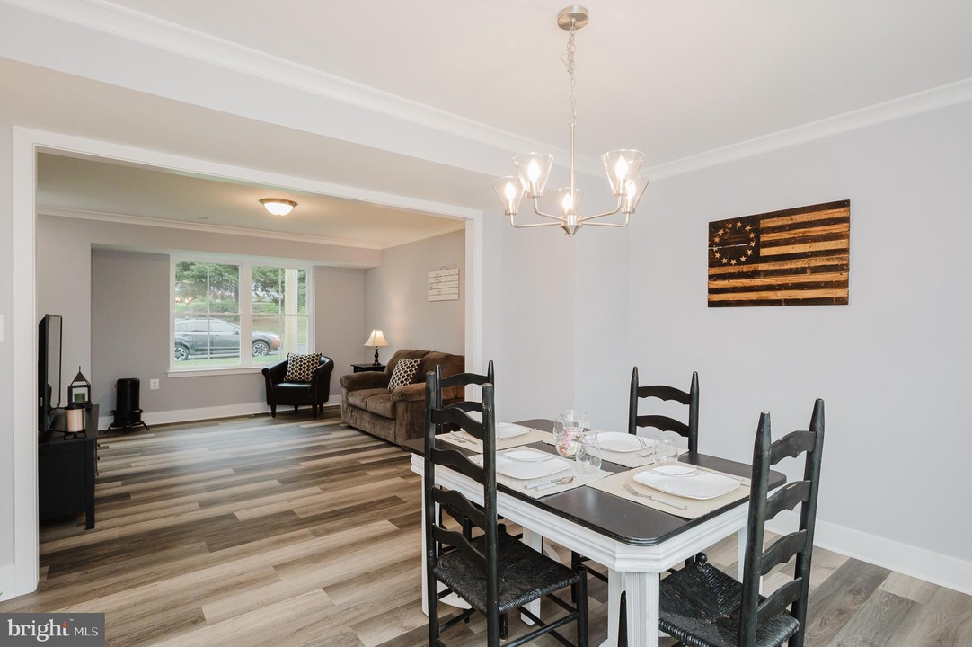 Open-concept dining area with a white table, black chairs, placemats, and a pendant chandelier; living room beyond.