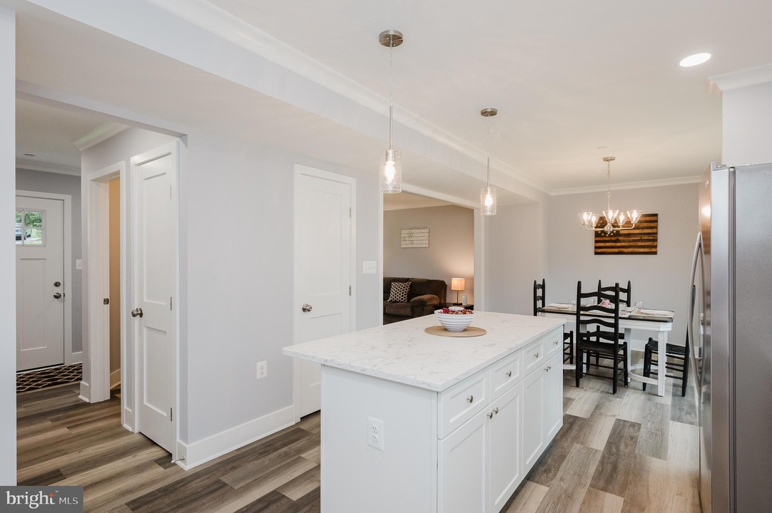 Open-concept kitchen with white island and marble countertop, pendant lights, and a dining area with black chairs in the background.