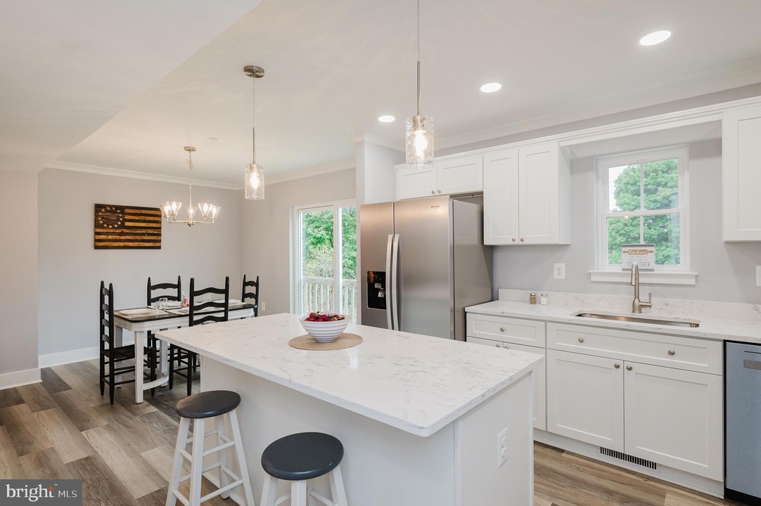 Bright modern kitchen with white cabinets, marble island, and stainless steel refrigerator; open dining area nearby with dark wooden chairs and a rectangular table.