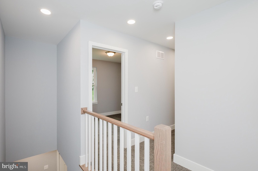 Bright upstairs hallway with gray walls, white trim, a wooden handrail and white balusters overlooking a staircase, and an open doorway to a room with a window.