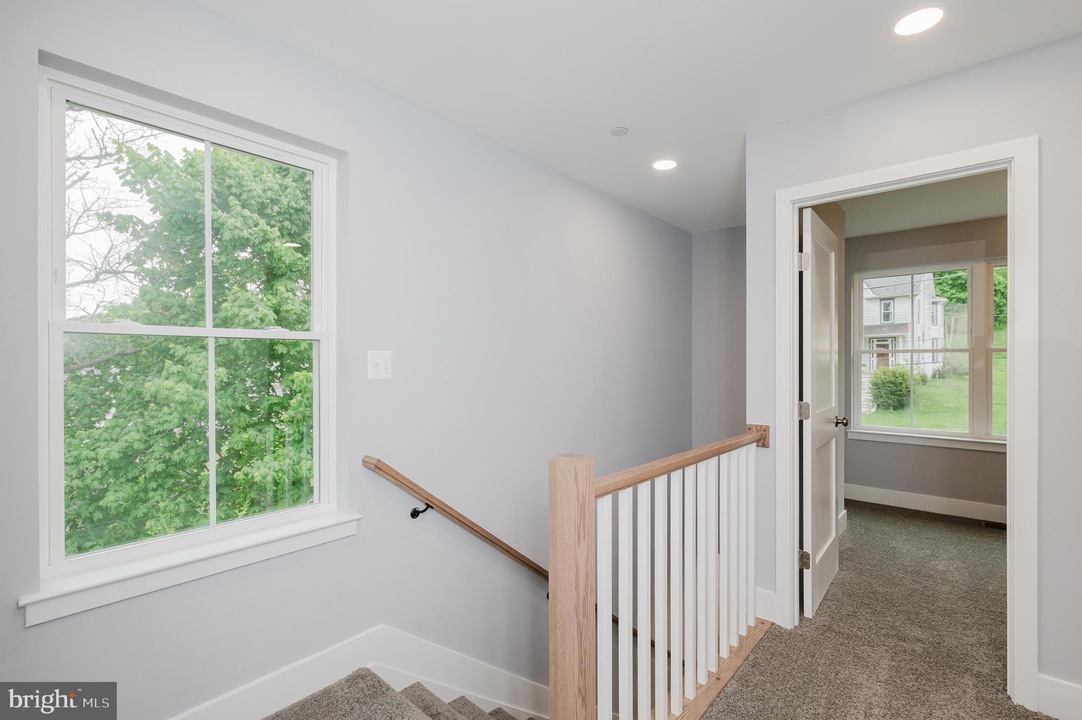 Interior stair landing with gray walls, carpeted stairs, and a wooden handrail leading down; bright window shows green trees outside and a doorway to an adjoining room.