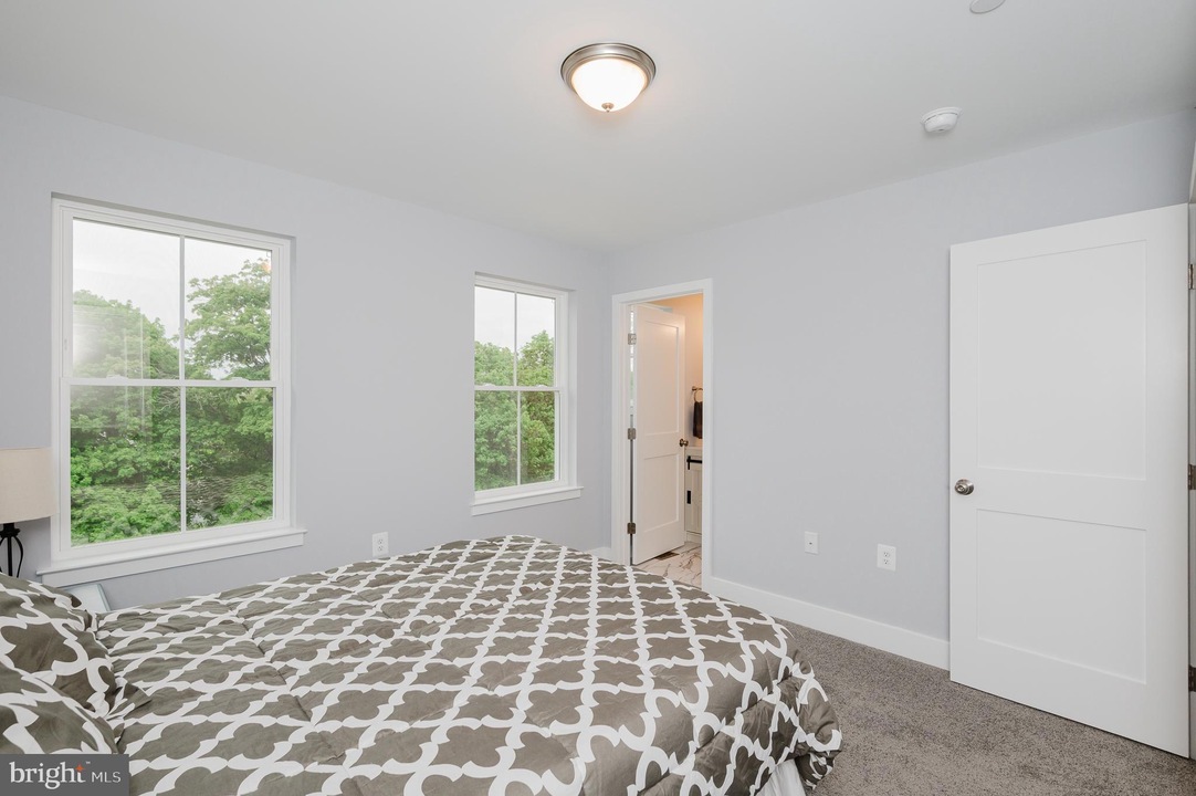 Bright bedroom with light gray walls, two large windows overlooking green trees, a bed with a gray-and-white patterned duvet, and a doorway to an en-suite bathroom.