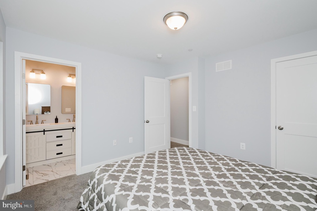 Bedroom with a gray-patterned comforter, light gray walls, and an open doorway to a white-turnished bathroom with double sinks and mirrors on marble floor.
