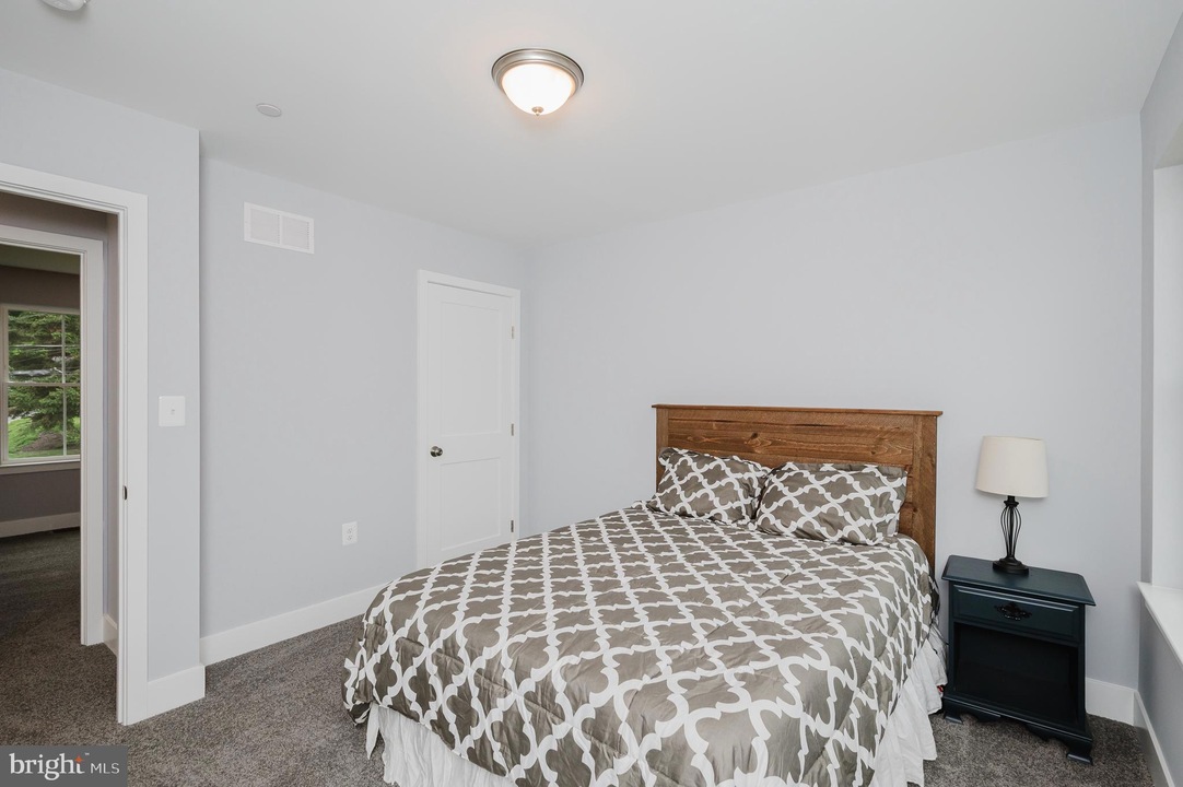 Bedroom with a wooden headboard bed, gray geometric bedding, two pillows, a dark nightstand with lamp, and pale gray walls.