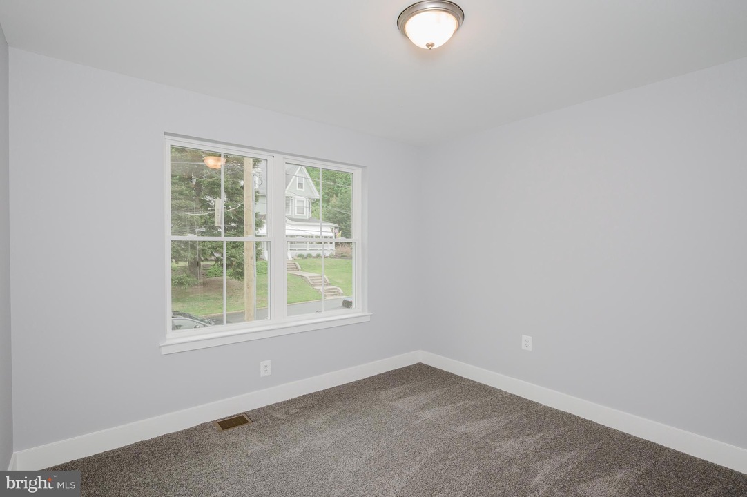 Empty bedroom with light gray walls, beige carpet, and a central ceiling light fixture; window shows a suburban street outside.