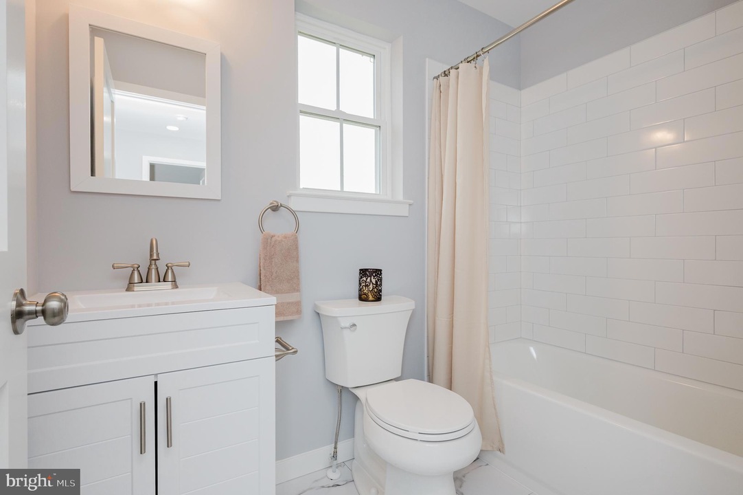 Bright white bathroom with a vanity, mirror, towel ring, toilet, and a bathtub with a beige curtain.