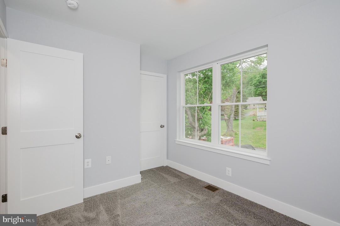 Empty bedroom with light gray walls, white doors, and a large window overlooking a green yard outside.