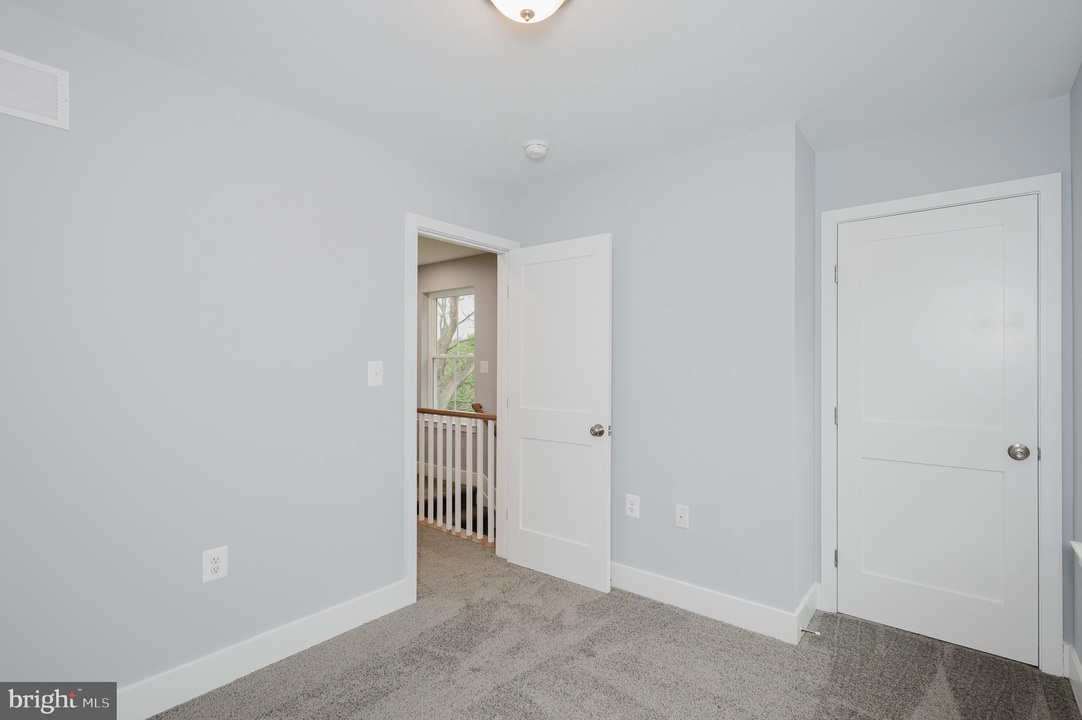 Empty light-blue bedroom with white doors, gray carpet, and baseboard trim; hallway visible beyond an open doorway.
