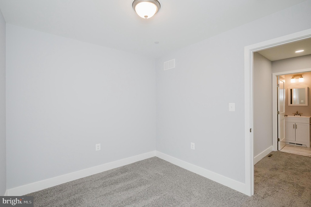 Empty small bedroom with light gray walls, white baseboards, carpet, ceiling light, and an open doorway to a bathroom.