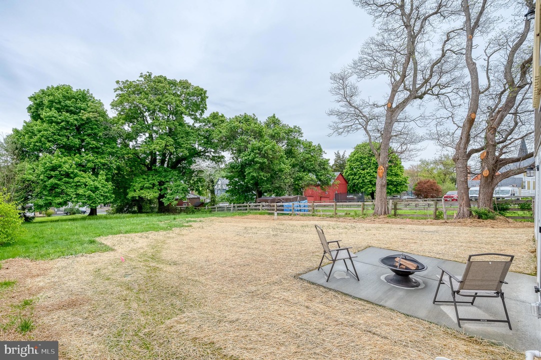 Backyard with a circular fire pit and three chairs on a concrete pad, straw-covered ground, and green trees in the background.
