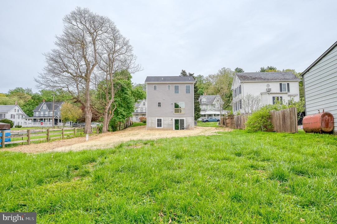 Backyard view with a three-story gray house in the center, a large bare tree on the left, and a wooden fence along the right edge; green lawn in the foreground.