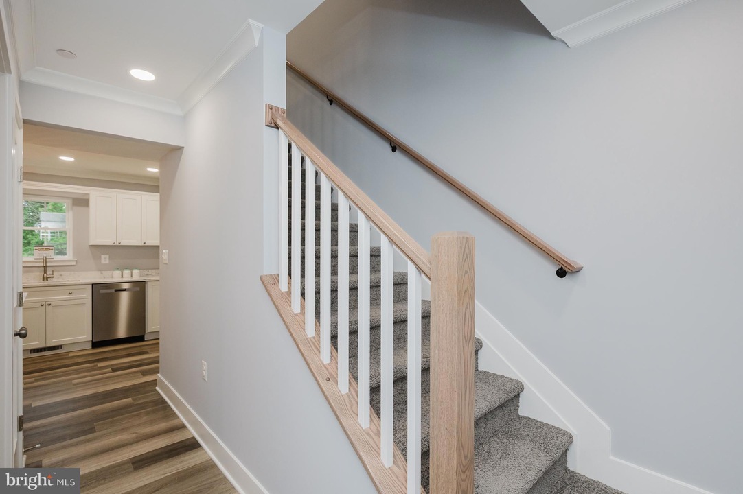 Staircase with light wood handrail and white balusters beside a hallway leading to a white kitchen with stainless appliances.