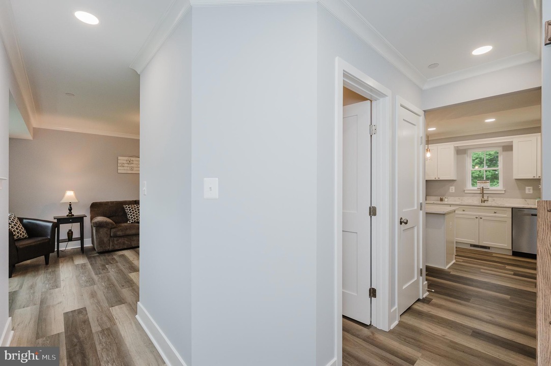 Bright hallway with light gray walls and wood-look flooring, opening to a living room on the left and a white kitchen on the right.