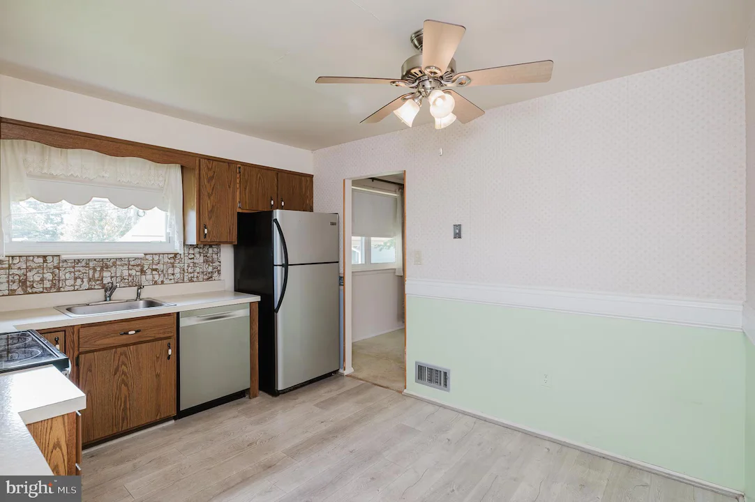 Kitchen with wooden cabinets, stainless steel fridge and dishwasher, double sink under a window, and a patterned backsplash behind the counters under a ceiling fan.