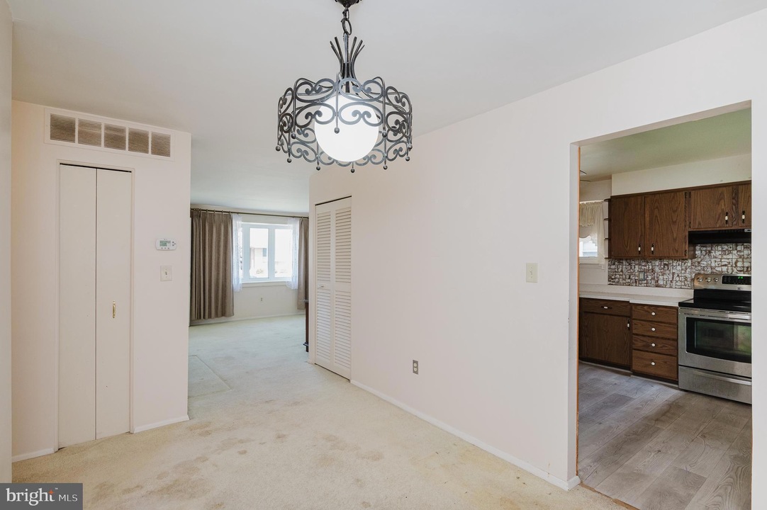 Living area with a decorative metal chandelier, cream carpet, and a doorway leading to a kitchen with wooden cabinets and stainless appliance.