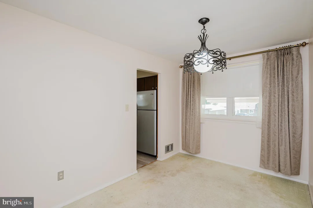 Empty living/dining room with beige walls, ornate black chandelier, and a window with taupe curtains; open doorway reveals a stainless steel refrigerator in the adjacent kitchen.