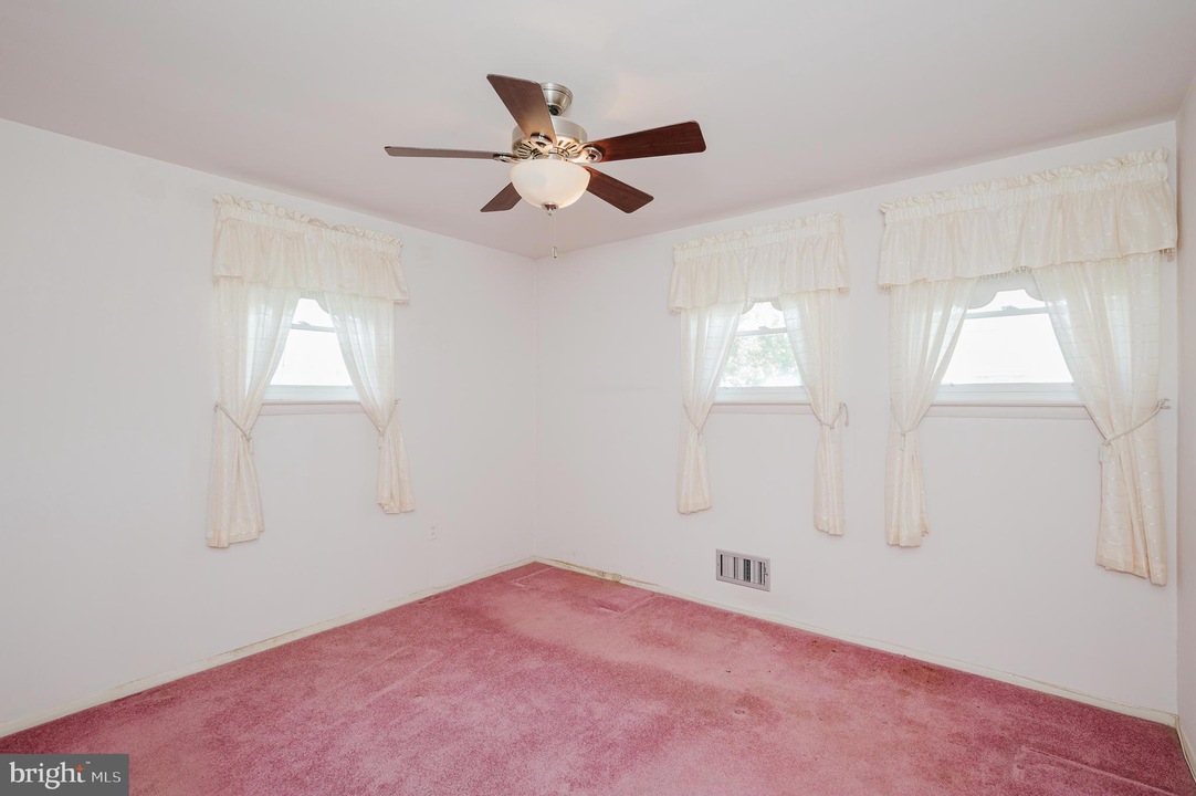Small empty bedroom with pink carpet, white walls, and three windows with lace curtains and a ceiling fan with lights.