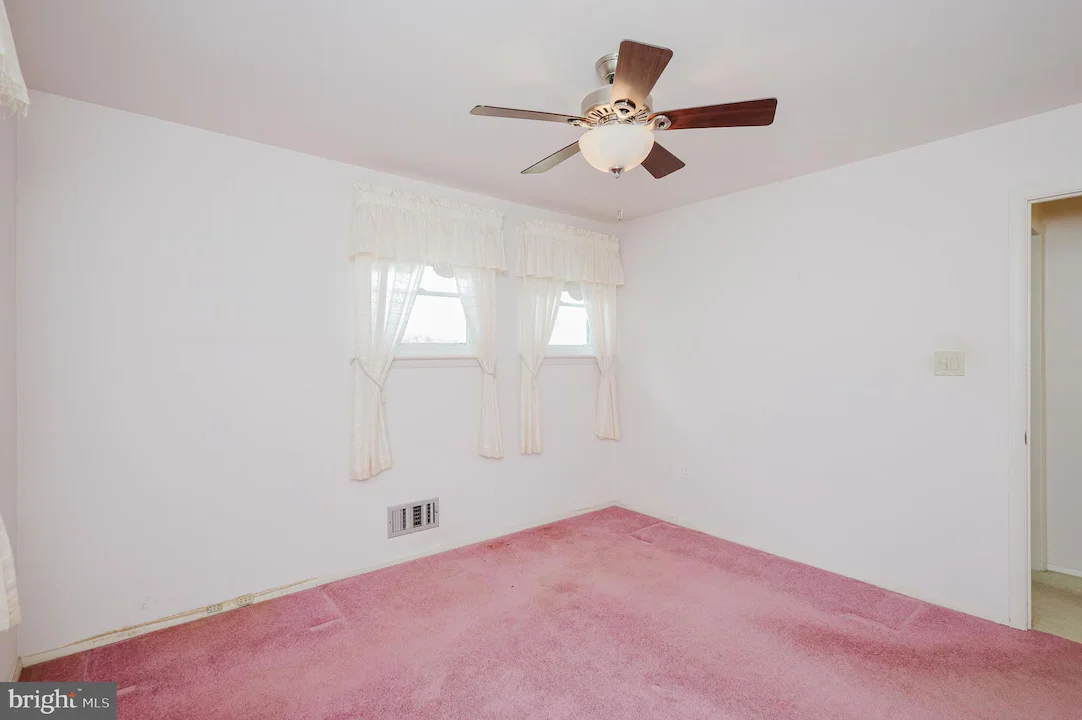Empty bedroom with white walls and pink carpet, a ceiling fan, two small windows with lace curtains, and a doorway to an adjoining room, plus a wall vent near the baseboard.