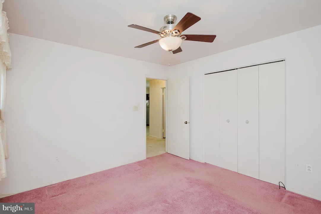 Empty bedroom with white walls, pink carpet, and a ceiling fan; large white bi-fold closet doors on one wall.