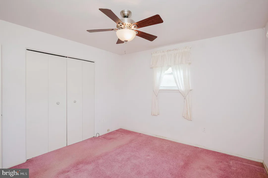 Empty bedroom with pink carpet, white walls, a ceiling fan, and a window with sheer curtains on the right.