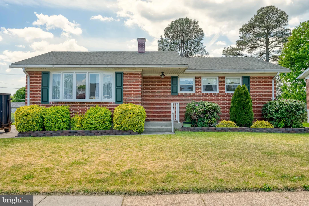 Brick single-story house with green shutters, a gray roof, and a row of trimmed boxwoods in the front yard.