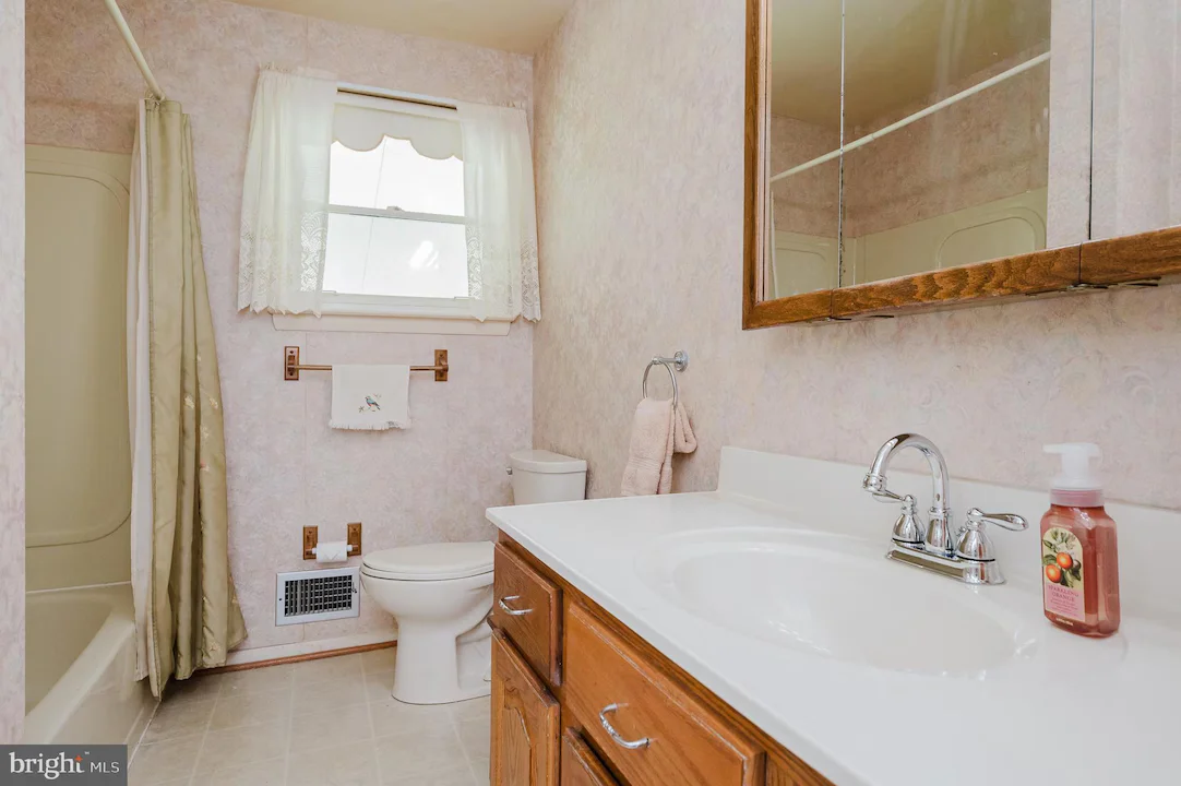 Bright pink bathroom with a white countertop sink, wooden vanity, chrome faucet, toilet, shower with a light curtain, and a window with lace curtains.