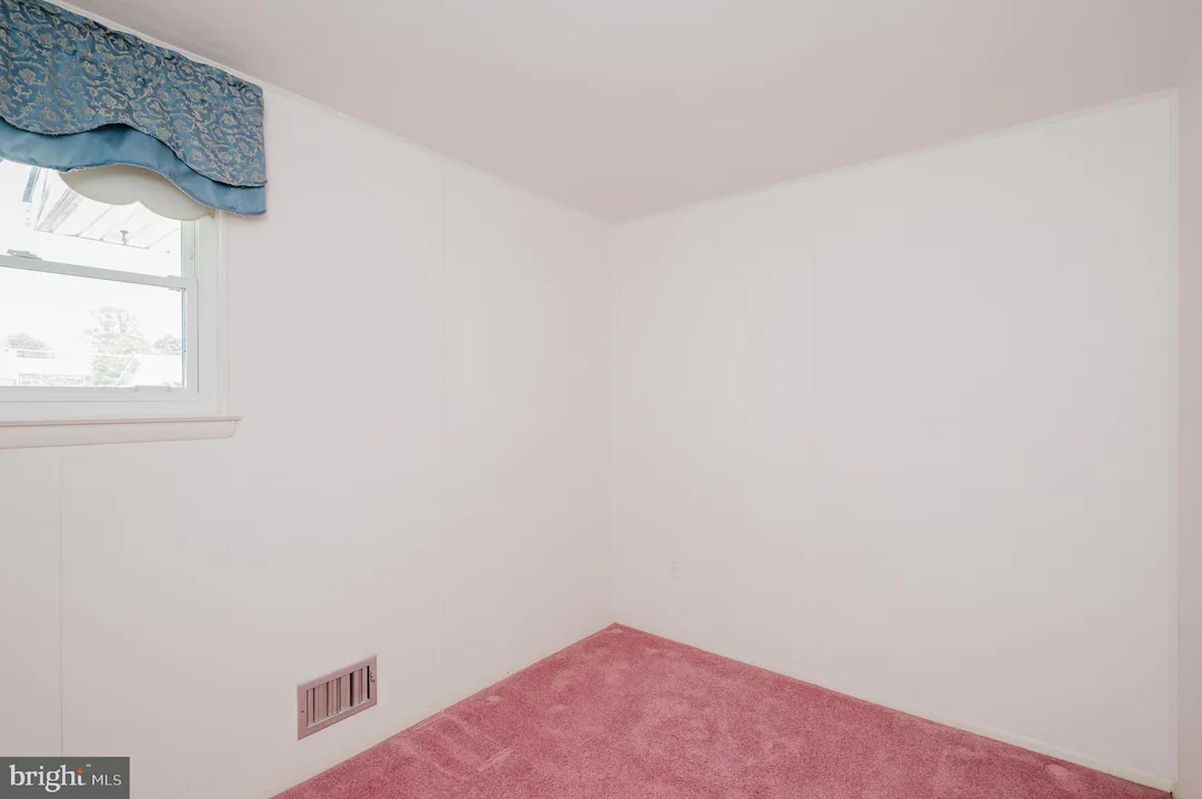 Empty corner of a small room with pink carpet, white walls, and a window with a blue valance.