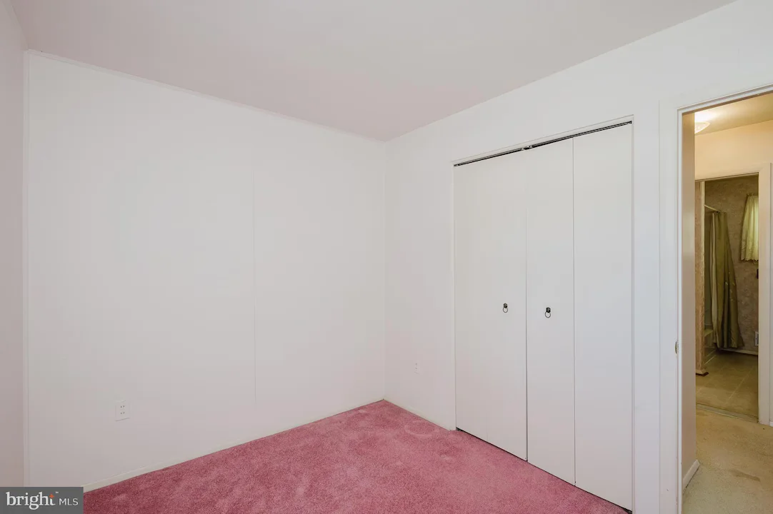 Empty bedroom with white walls, pink carpet, and a double-door closet; doorway to a bathroom is visible on the right.
