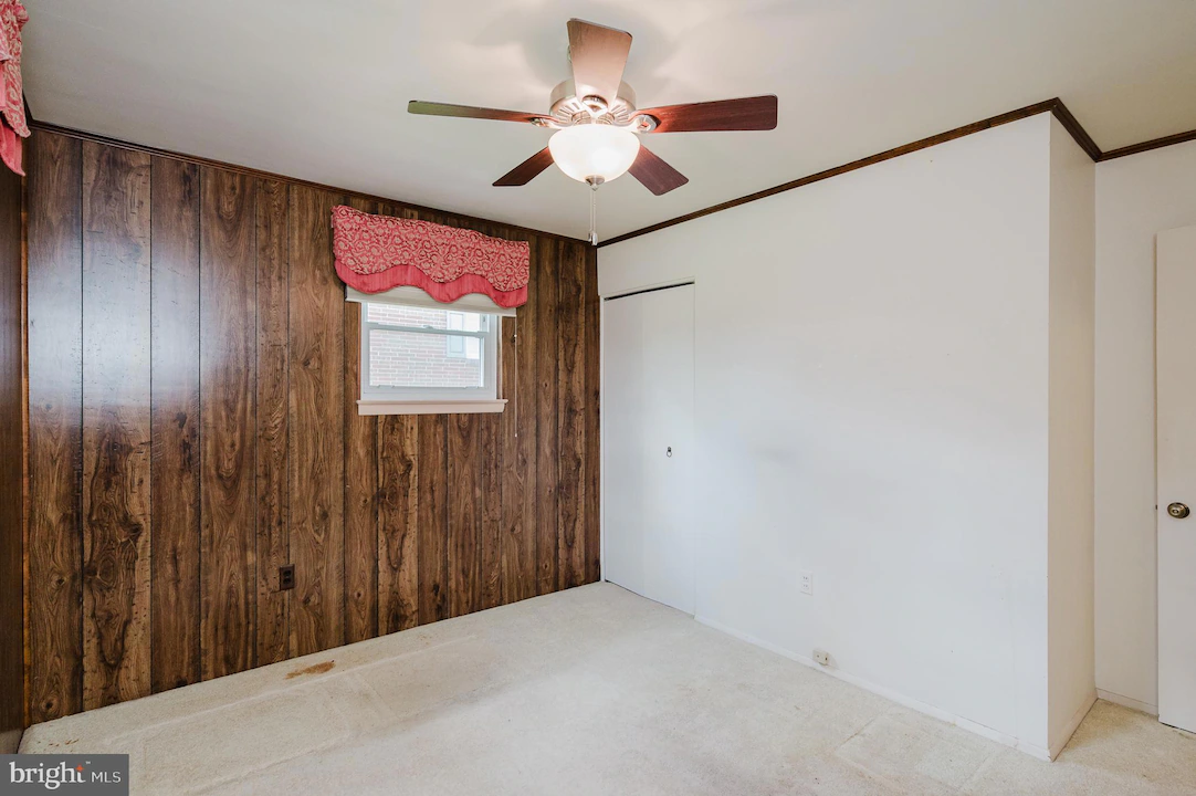 Room with a wood-paneled accent wall, small window with pink valance, ceiling fan, and white walls; beige carpet floor, closet door on the back wall.