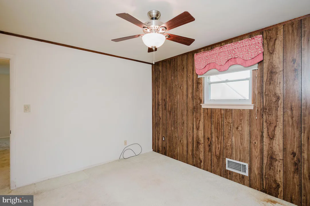 Empty room with white walls and a wood-paneled accent wall, featuring a ceiling fan with lights and a small window with a pink valance.