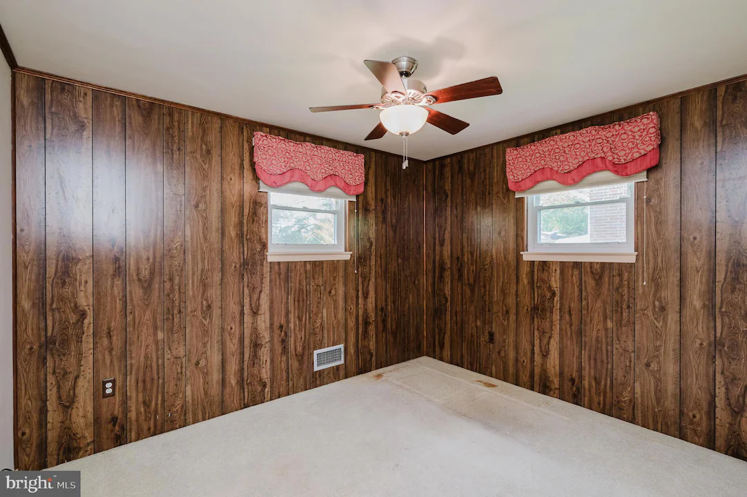 Basement-style room with dark wood paneling, two small windows with pink valances, a ceiling fan, and a stained beige carpet.