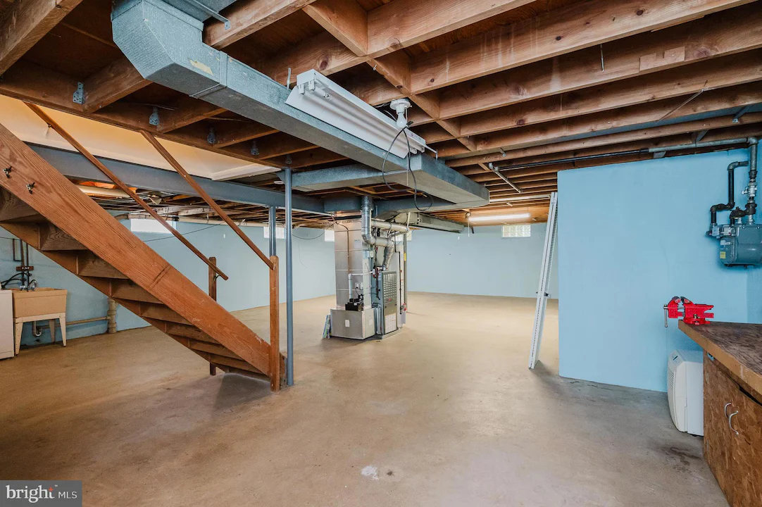Unfinished basement with wooden stairs, exposed beams, and utility equipment along a blue wall, concrete floor, and a workbench on the right side.
