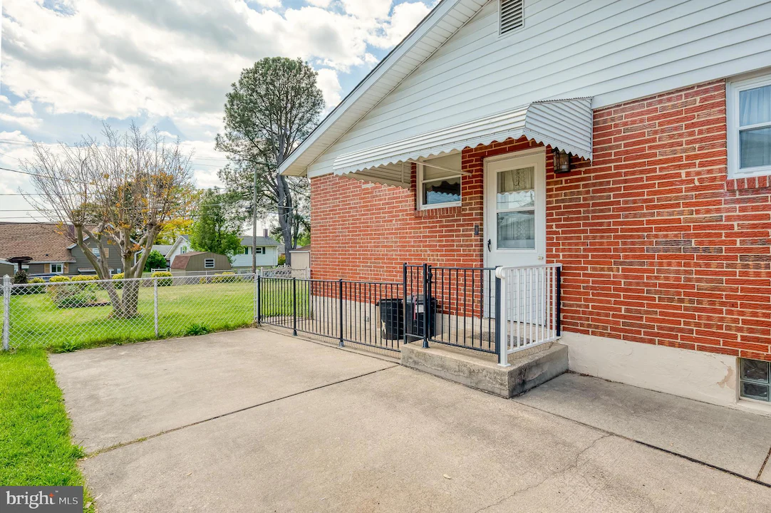 Brick house with small front porch, white door, and black railing; concrete driveway and chain-link fence in a suburban yard.
