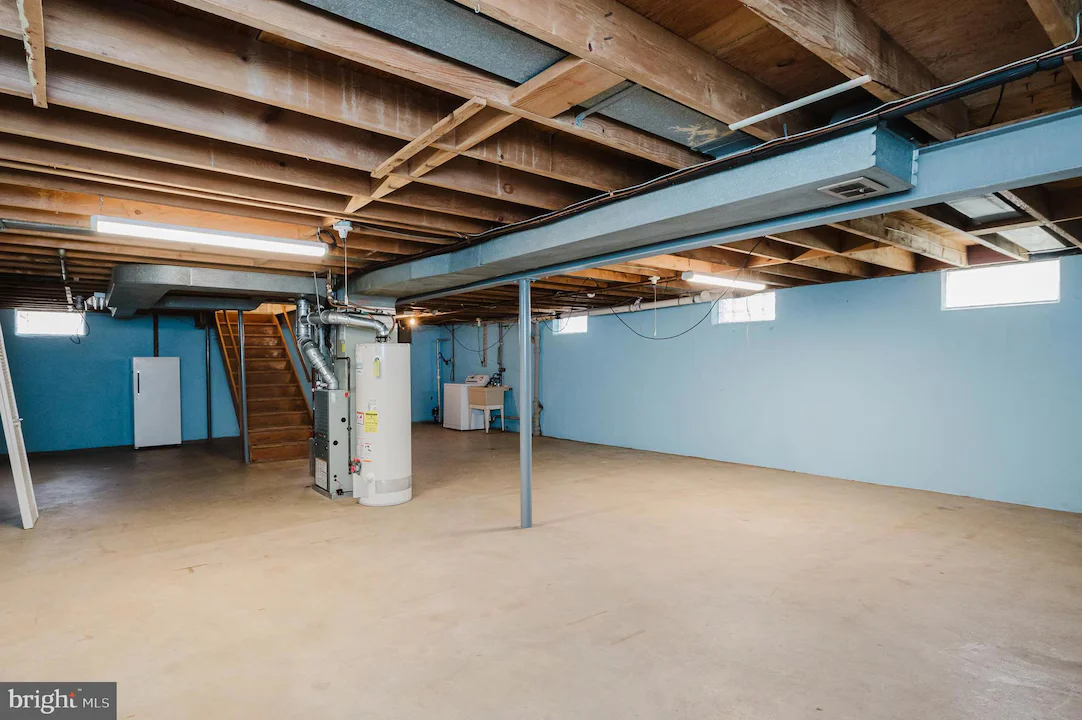 Unfinished basement with blue walls, exposed ceiling joists, a water heater and furnace, and a wooden staircase.