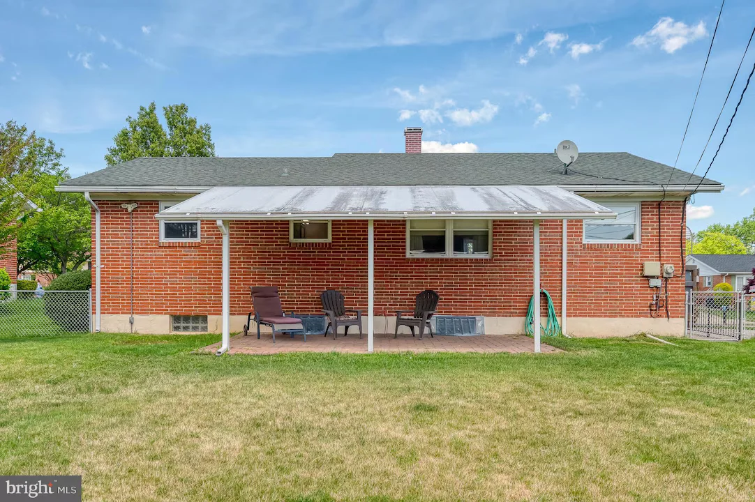 Brick house rear with a covered patio; four lawn chairs and a small table on a brick floor, green lawn beyond.