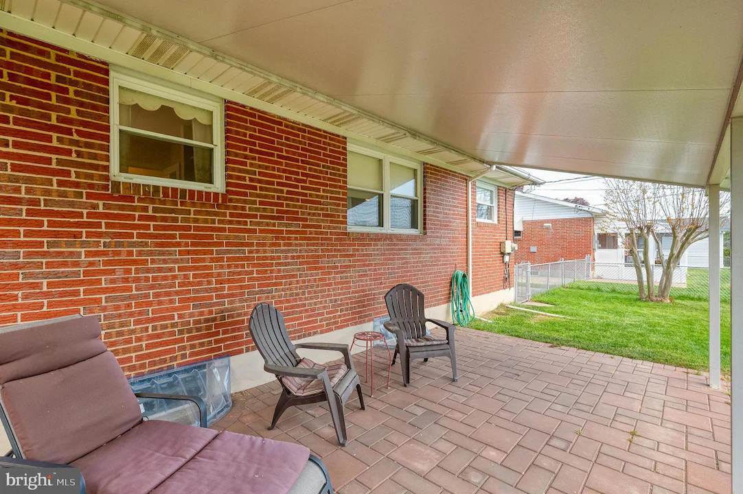 Covered brick-backyard patio with brown cushioned lounge chair and two plastic chairs on a paved surface; a green hose coiled on the wall and a grassy yard beyond a chain-link fence.