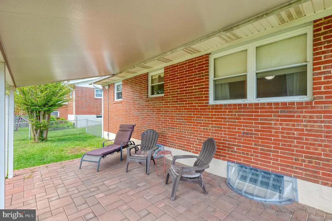 Covered brick patio with four outdoor chairs and a lounge chair along a red brick house, facing a fenced yard.