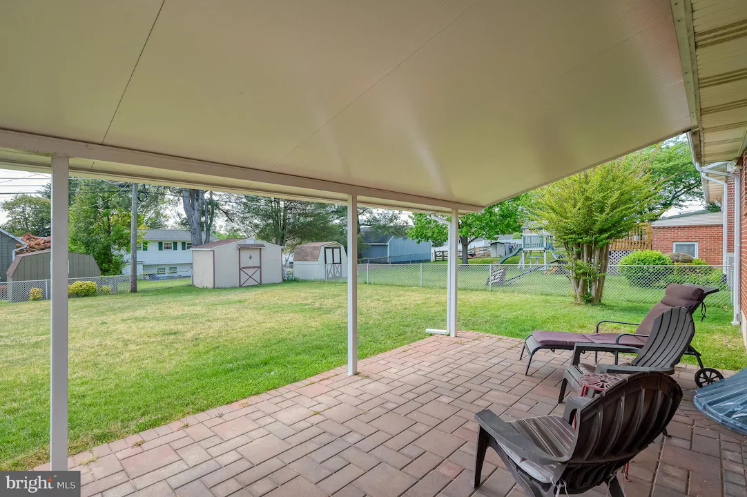 Covered patio overlooking a green backyard with lawn, sheds, and a chain-link fence; outdoor chairs and a lounge chair on brick patio.