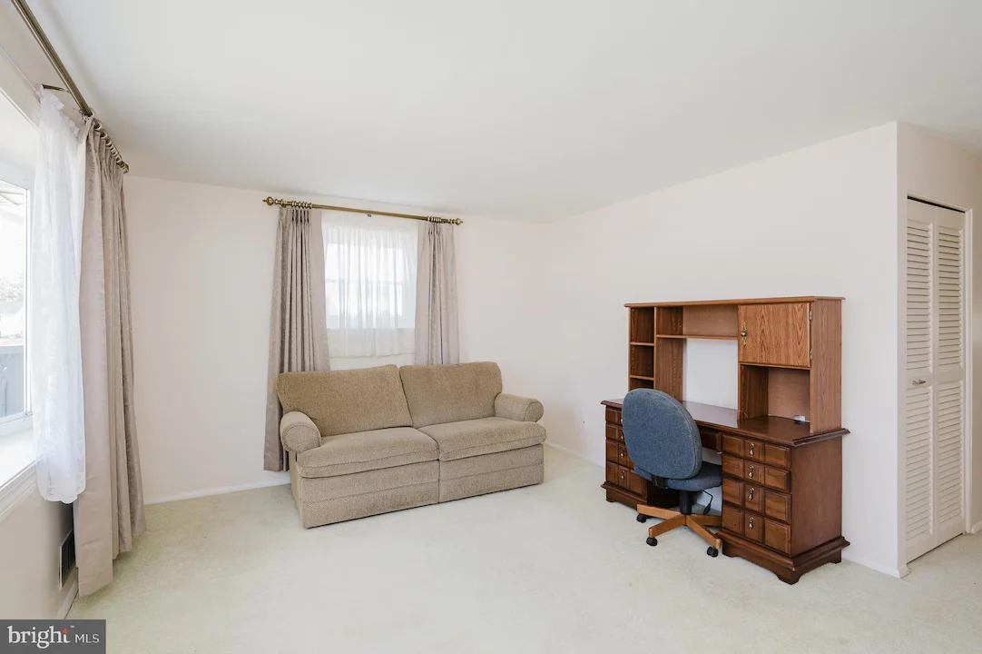 A small living room with a beige sofa, light walls, and a wooden desk with a blue rolling chair against the right wall, under a window with gray curtains.