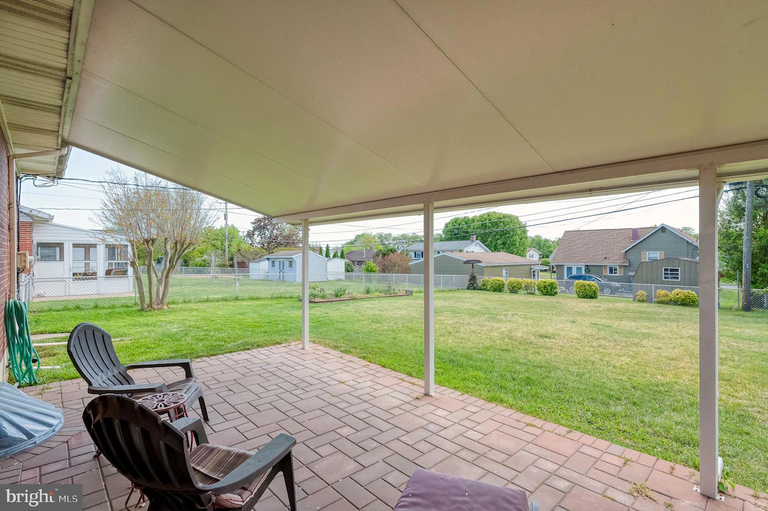 Covered patio with brick pavers and three black plastic chairs facing a green backyard with a chain-link fence and neighboring houses in the distance.
