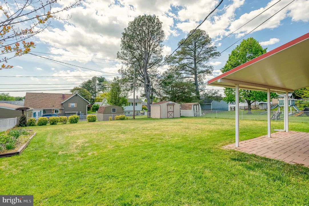 Wide view of a suburban backyard with a green lawn, sheds, and tall trees beyond a chain-link fence, plus a covered patio on the right.