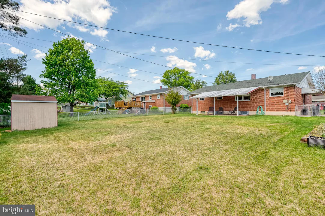 Spacious backyard with a brick house, covered patio, and a wooden playset; a shed sits near a chain-link fence under a bright sky.